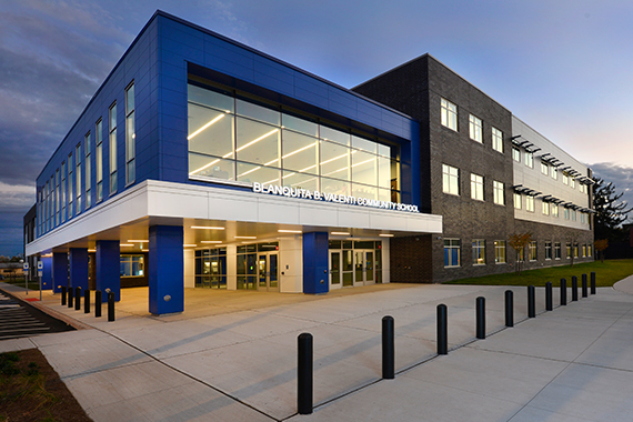 An exterior view of the Blanquita B. Valenti Community School in New Brunswick, NJ shows the main entrance and security bollards. The second floor, elevated on columns, looks into the large glass windows that are a centerpiece of the media center.