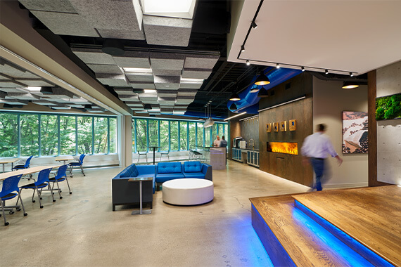 One of the staff lounges at the administrative offices of Blue Foundry Bank, showing colleagues having coffee next to a fire feature. Partitions, separating the adjacent training center, are open. In the foreground, a stage used for company-wide presentations is seen.